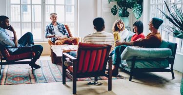 A small group of adults talks together while seated on chairs and a couch. Large windows let in ample natural light.