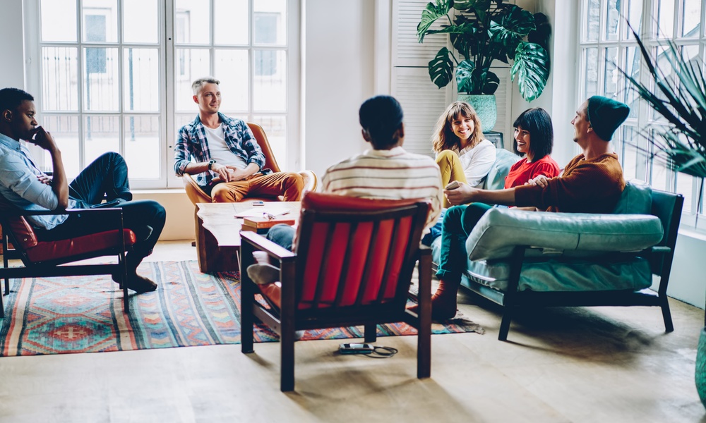 A small group of adults talks together while seated on chairs and a couch. Large windows let in ample natural light.