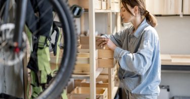 A woman organizes items on wooden storage shelves in a garage, with a bicycle wheel and hanging gear nearby.