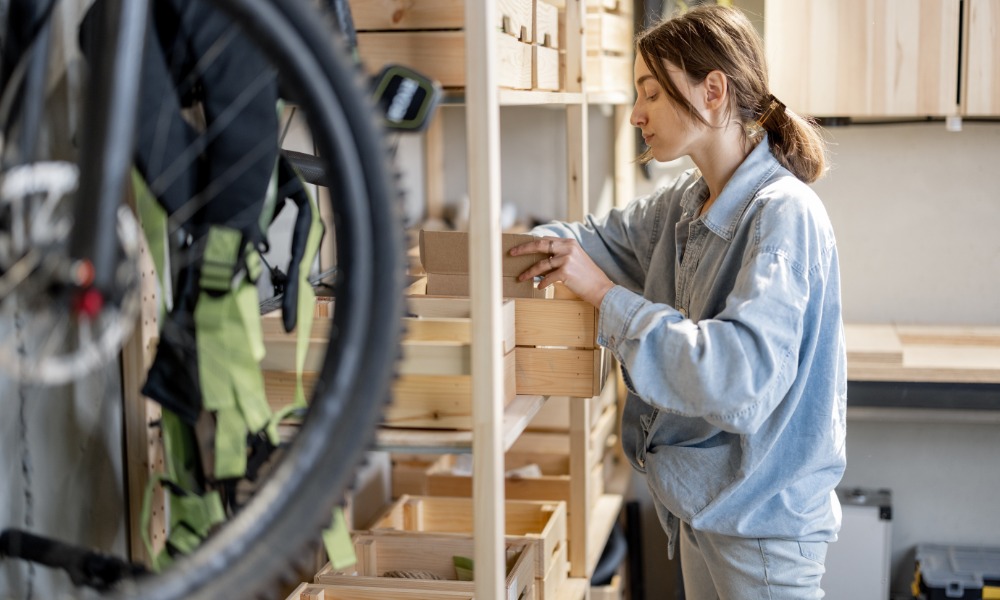 A woman organizes items on wooden storage shelves in a garage, with a bicycle wheel and hanging gear nearby.