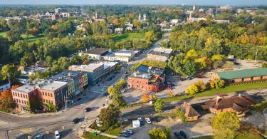 Aerial view of a small town with colorful downtown buildings, tree-lined streets, parked cars, and surrounding greenery.