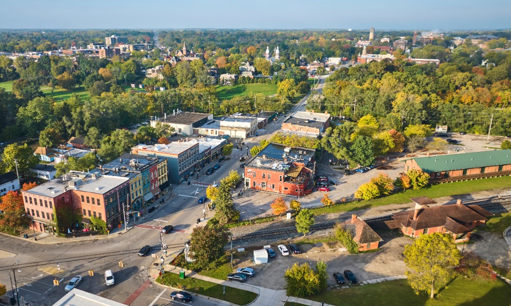 Aerial view of a small town with colorful downtown buildings, tree-lined streets, parked cars, and surrounding greenery.