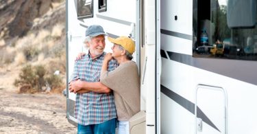 An older couple smiles and leans against the doorway of a white motorhome parked beside a rocky desert landscape.