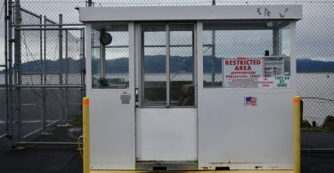 A small white security booth outside near a large lake. A red sign in the booth window reads “Restricted Area”.