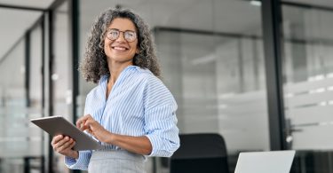 A professional wearing glasses smiles and stands near windows in an office. The professional holds a tablet.