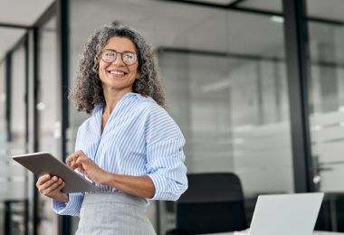 A professional wearing glasses smiles and stands near windows in an office. The professional holds a tablet.