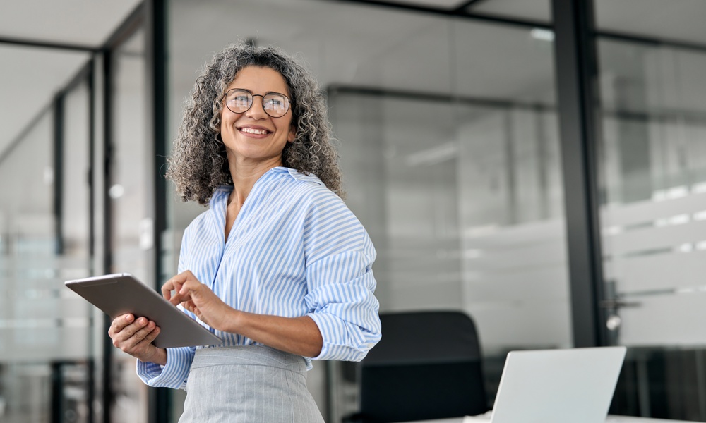 A professional wearing glasses smiles and stands near windows in an office. The professional holds a tablet.