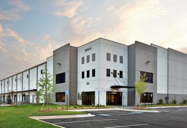 Exterior of a large modern industrial warehouse with loading docks, office entrances, landscaped grounds, and an empty parking lot.