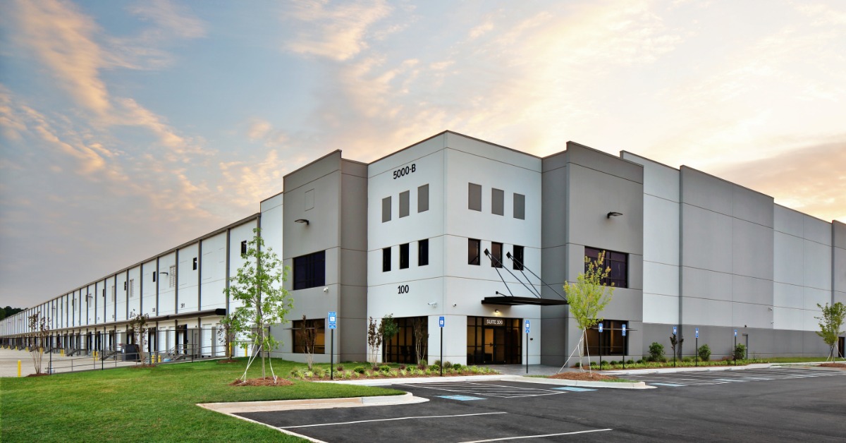 Exterior of a large modern industrial warehouse with loading docks, office entrances, landscaped grounds, and an empty parking lot.